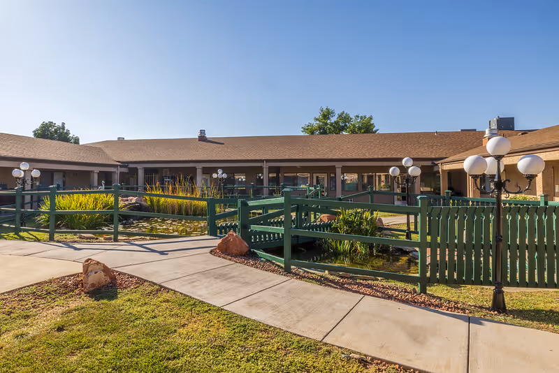 Outdoor courtyard area of Verde Valley Assisted Living featuring a green wooden fence, a small pond with aquatic plants, multiple lamp posts with round white lights, and a single-story building with a brown roof in the background under a clear blue sky.