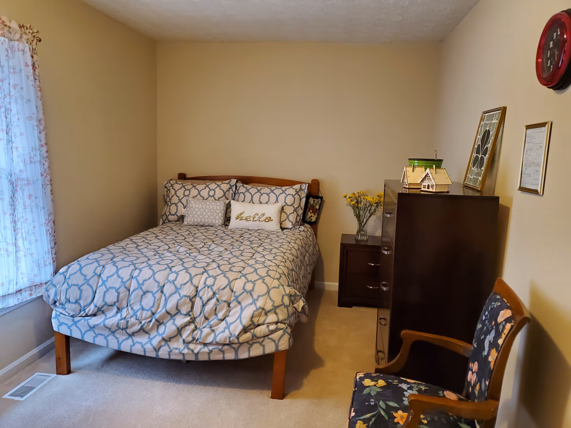 Small furnished bedroom with a patterned bed against a beige wall, a dark dresser and nightstand, and a floral armchair.
