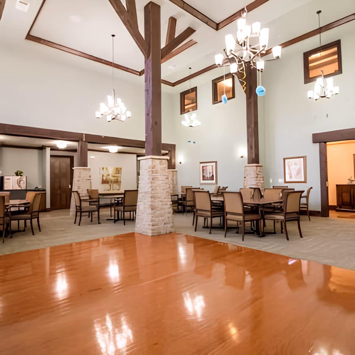 Spacious senior-living common dining area with high ceilings, wooden beams, chandeliers, and multiple tables and chairs.