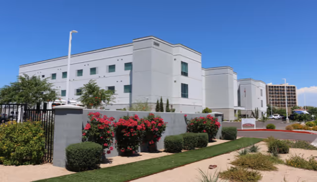 Exterior view of a modern three-story healthcare facility building with white and gray walls, multiple windows, and a landscaped area with bushes and red flowering plants in front. The sky is clear and blue.
