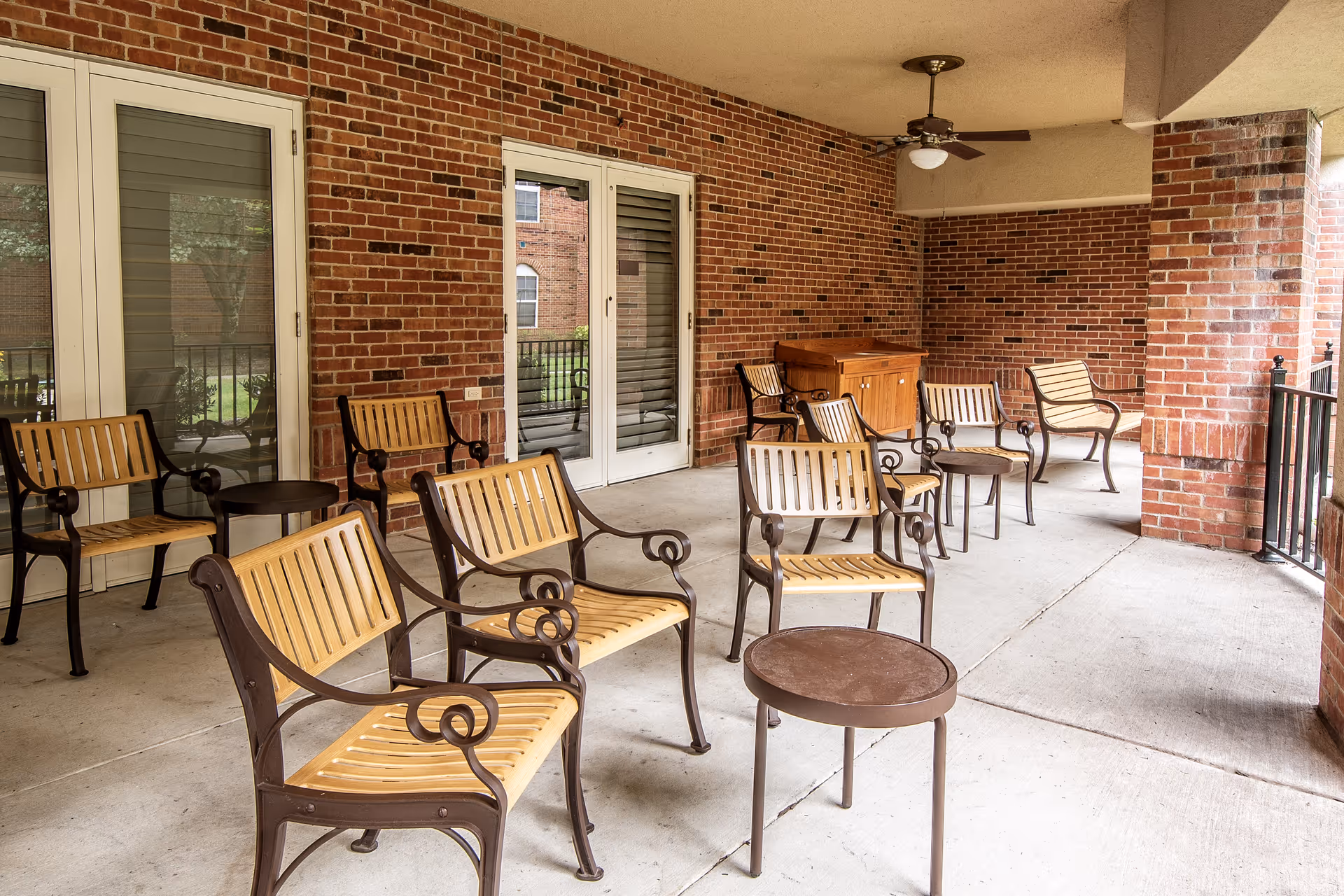 Covered outdoor patio area with multiple wooden and metal chairs arranged around small round tables. The patio has brick walls, a ceiling fan, and glass doors leading inside. There is a wooden cabinet against the far wall.
