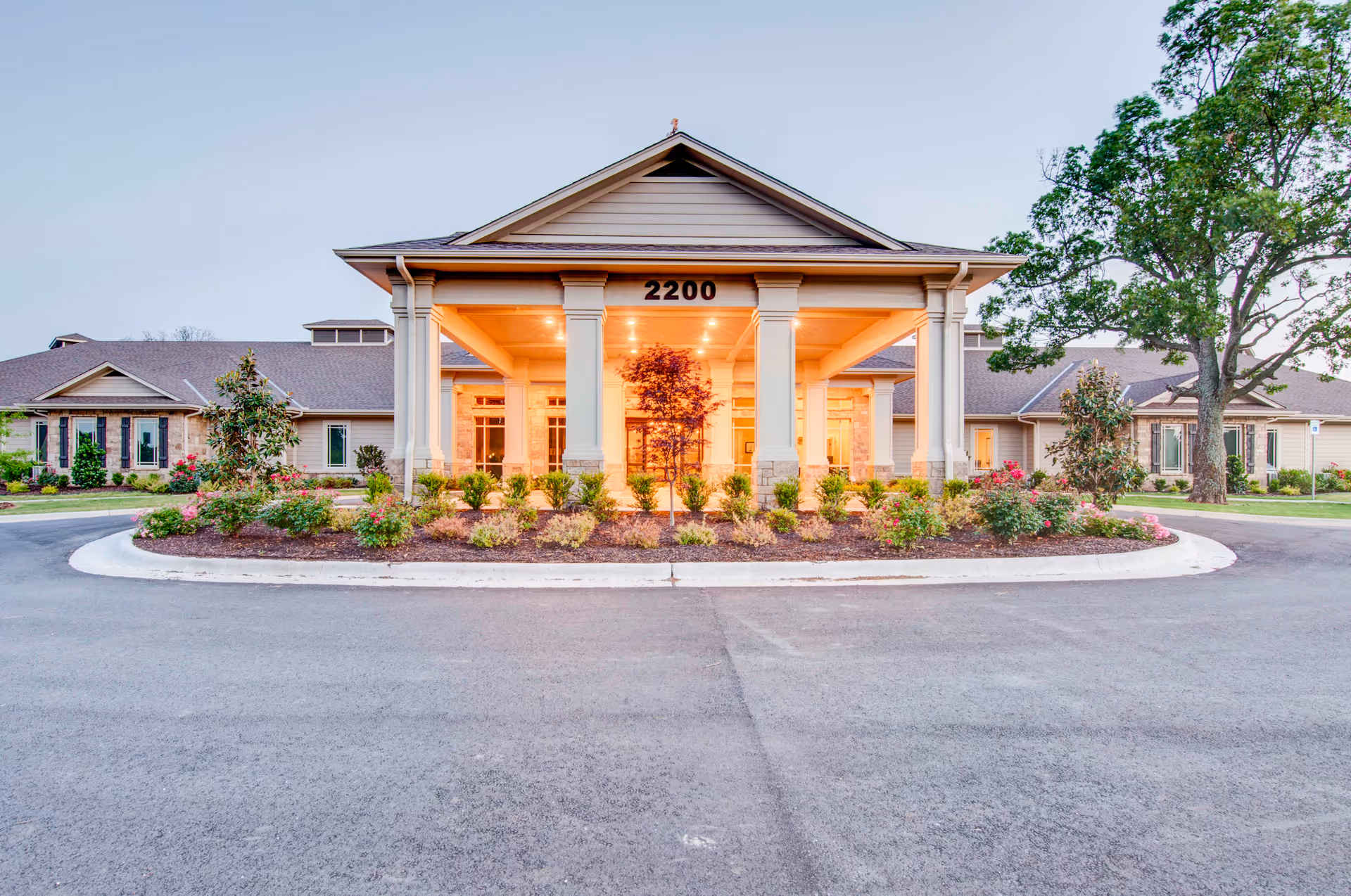 Front exterior view of Innisfree Nursing Center building with a covered entrance supported by columns, illuminated warmly from inside. The building is surrounded by landscaped bushes, flowers, and trees, with a paved driveway in front.