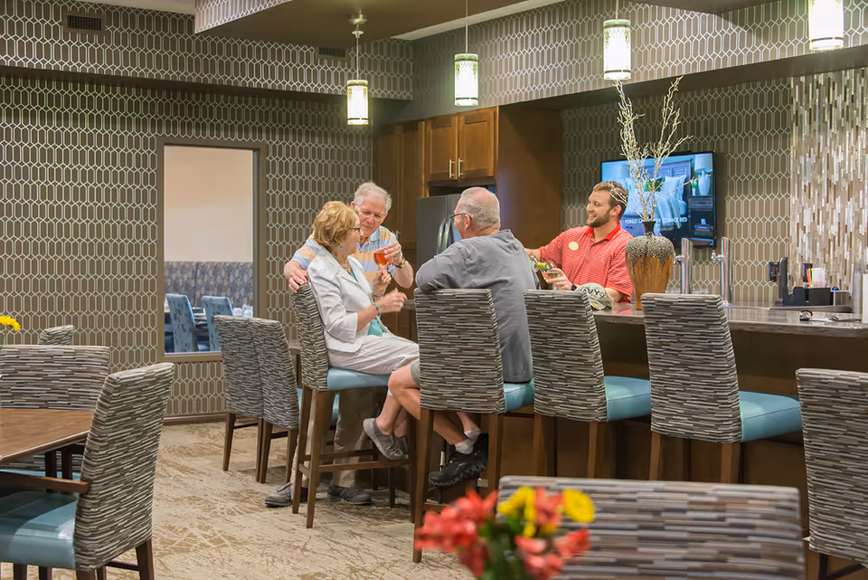 Three elderly people sitting on bar stools at a kitchen island counter, enjoying drinks and conversation with a younger man in a red shirt standing behind the counter. The room has patterned wallpaper, pendant lights, and a TV mounted on the wall.