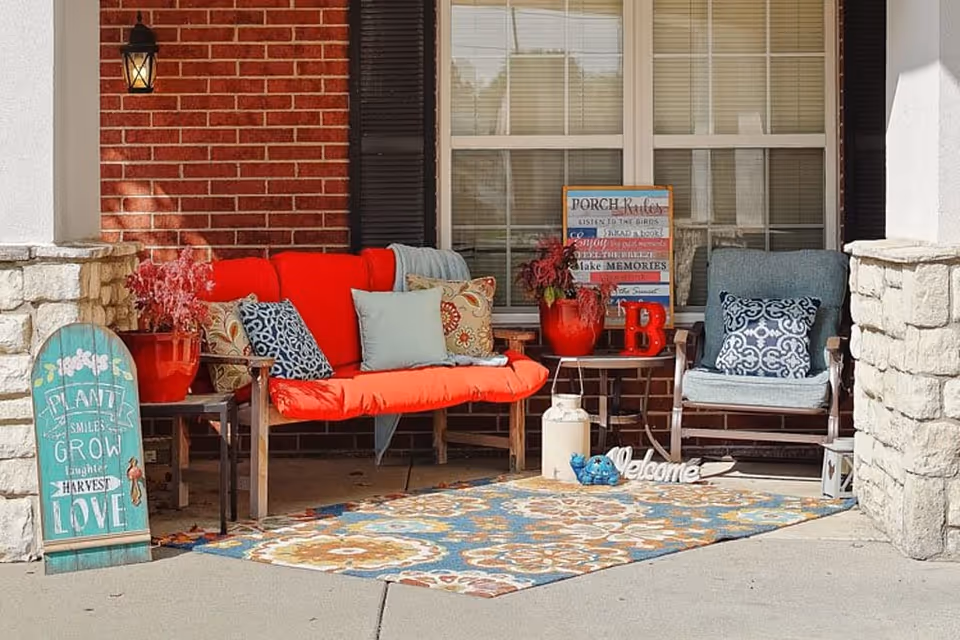 A cozy front porch area with a red cushioned bench and a blue cushioned chair, both adorned with decorative pillows. There is a colorful patterned outdoor rug on the concrete floor. A small table between the seating holds red flower pots and decorative items, including a sign with porch rules. A green wooden sign with inspirational words leans against a stone pillar on the left. The porch is framed by brick and stone walls with a window in the background.