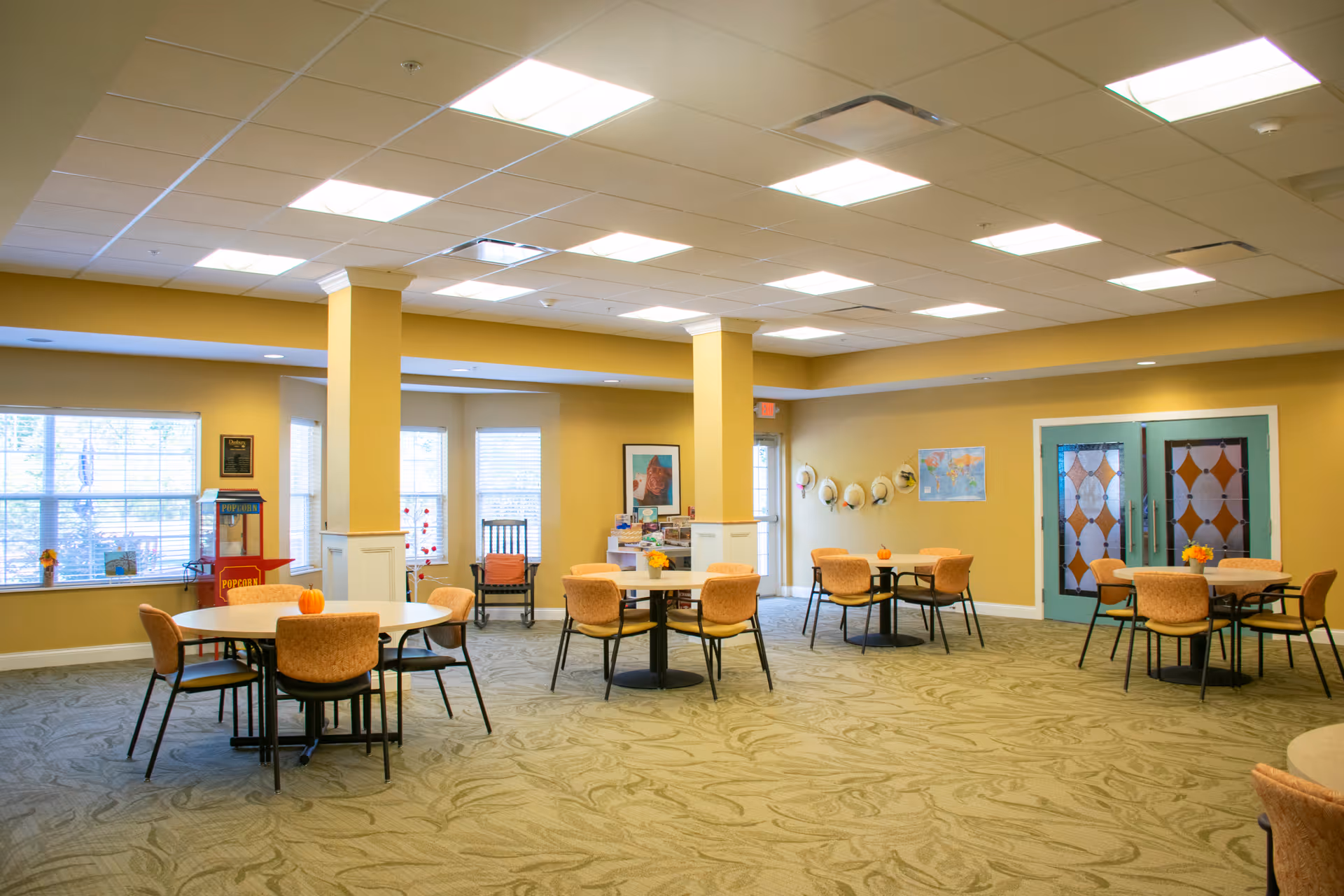 A spacious, well-lit common area with round tables and orange cushioned chairs arranged throughout the room. The walls are painted yellow with white trim, and there are large windows letting in natural light. Decorative items like small pumpkins on tables, a popcorn machine, framed artwork, and a row of hats hanging on the wall add a cozy touch. The room has a patterned carpet and ceiling lights.