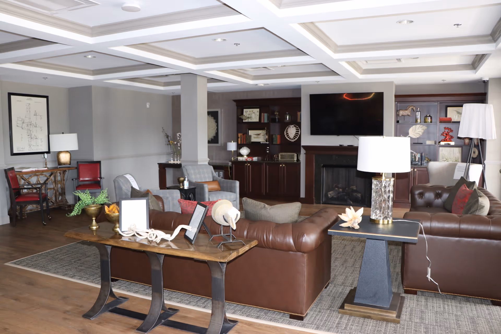 A spacious and well-lit living room featuring two brown leather sofas, two gray armchairs, a large flat-screen TV mounted above a fireplace, and wooden bookshelves with decorative items. The room has a coffered ceiling, wooden flooring, and various lamps and decor pieces on tables.