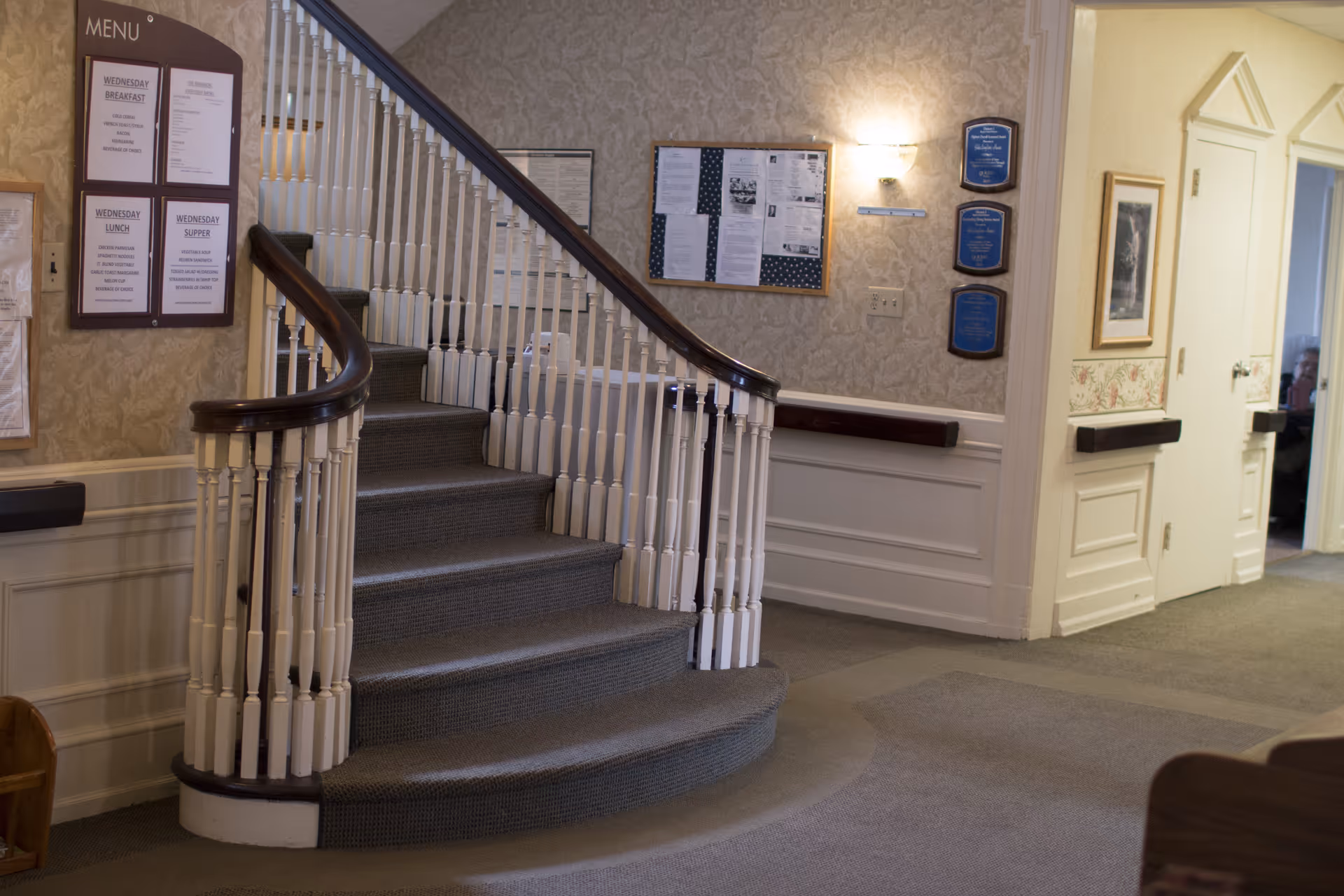 Interior view of a senior living facility showing a carpeted staircase with white spindles and dark wooden handrails. The walls are decorated with patterned wallpaper, bulletin boards, plaques, and a framed picture. There is a menu board on the left side of the staircase and a hallway with doors on the right.