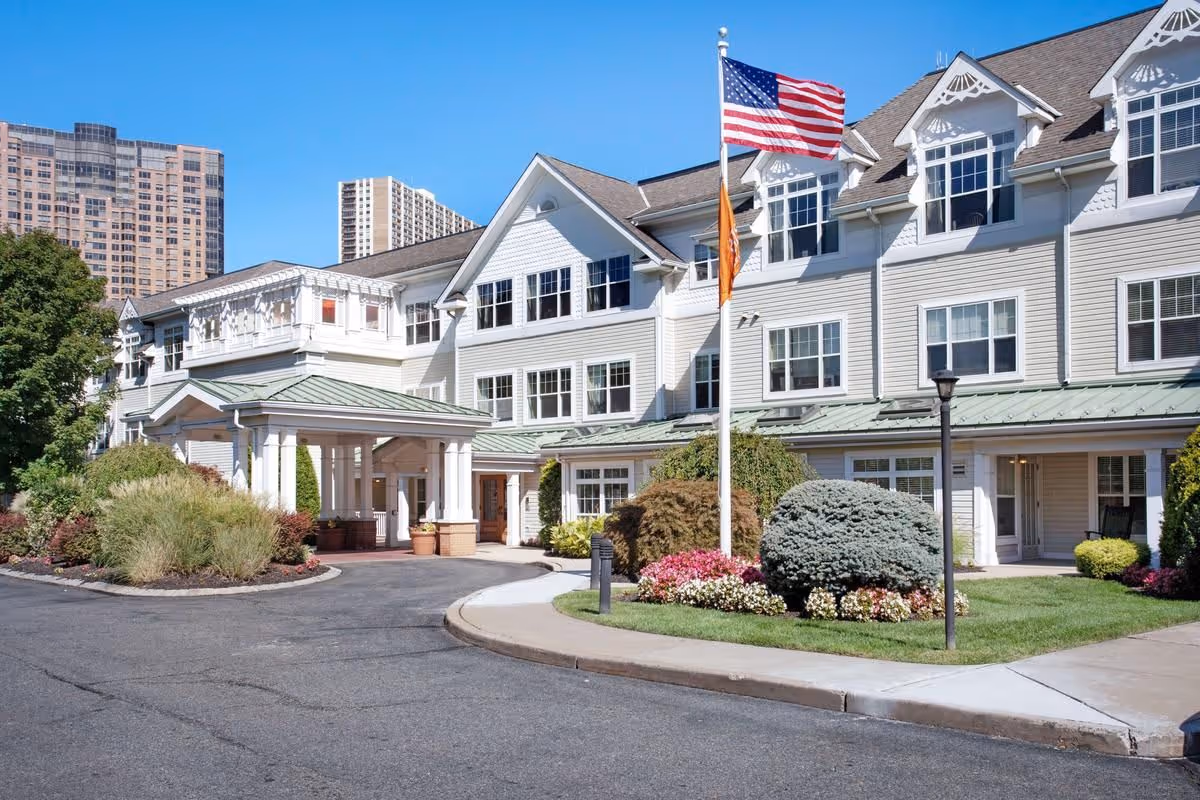 Exterior view of a senior living facility building with white siding and multiple windows, a covered entrance with a green roof, landscaped bushes and flowers, and an American flag on a flagpole in front under a clear blue sky.