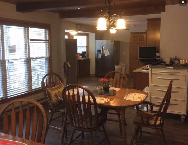 A dining area with a wooden table and six wooden chairs, some with cushions. The table has placemats and a small plant centerpiece. Large windows with blinds are on the left side, and a kitchen area with a refrigerator, cabinets, and a countertop is visible in the background. A ceiling light fixture with three bulbs hangs above the table.