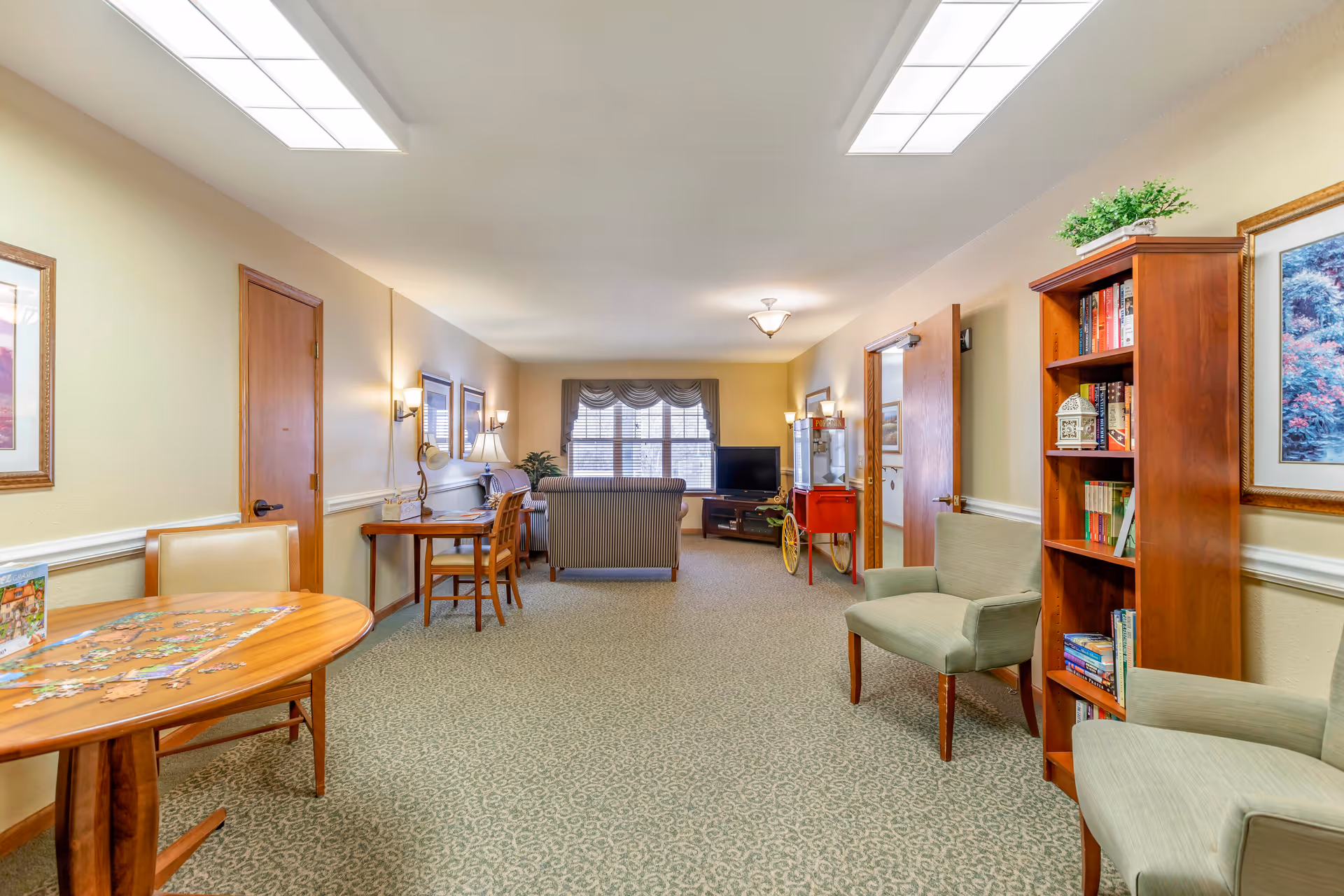 A bright communal living room in a senior facility with seating, a TV, bookshelves, and a table with a puzzle.