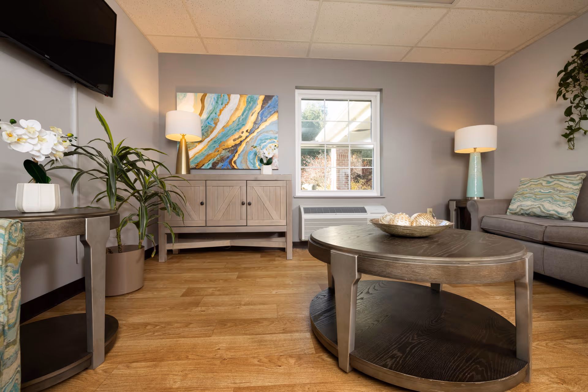 A cozy living room in a senior care facility featuring a gray sofa with patterned cushions, a wooden oval coffee table with decorative items, a wooden sideboard with a colorful abstract painting above it, two table lamps, a flat-screen TV mounted on the wall, and several potted plants. The room has light gray walls and a window letting in natural light.