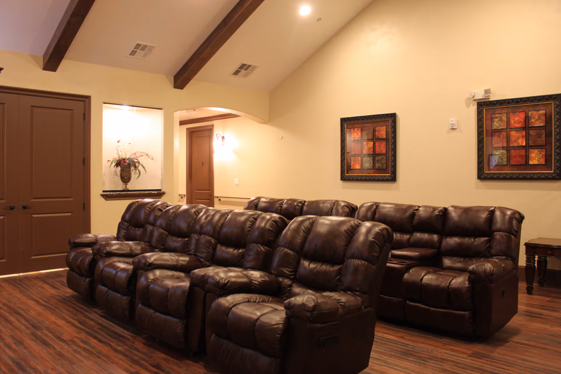 A cozy seating area with multiple dark brown leather recliners arranged in two rows facing forward. The room has wooden flooring, beige walls, and a vaulted ceiling with exposed wooden beams. Two framed abstract art pieces hang on the wall, and there is a small side table to the right. A double door and an alcove with a decorative vase are visible in the background.