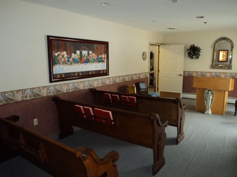 Small chapel-style interior with wooden pews facing a lectern and religious artwork on the walls.