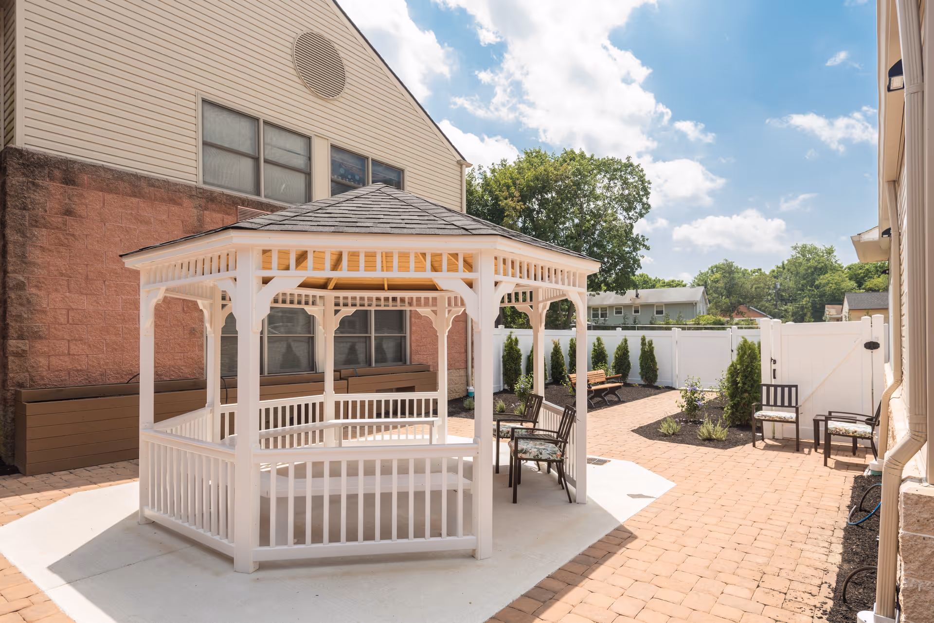Outdoor courtyard area with a white wooden gazebo in the center, surrounded by brick paving. There are several chairs and benches placed around the courtyard, with a white fence and some greenery in the background under a partly cloudy sky.