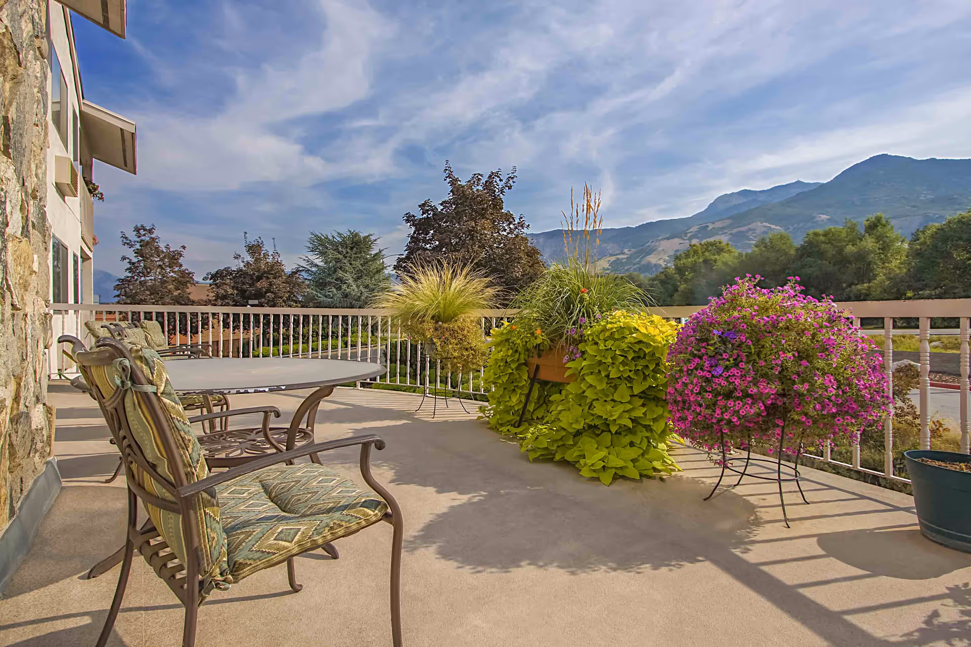 Outdoor patio area with cushioned metal chairs and a round table, surrounded by potted plants with green and purple foliage. In the background, there are trees and mountains under a partly cloudy sky.