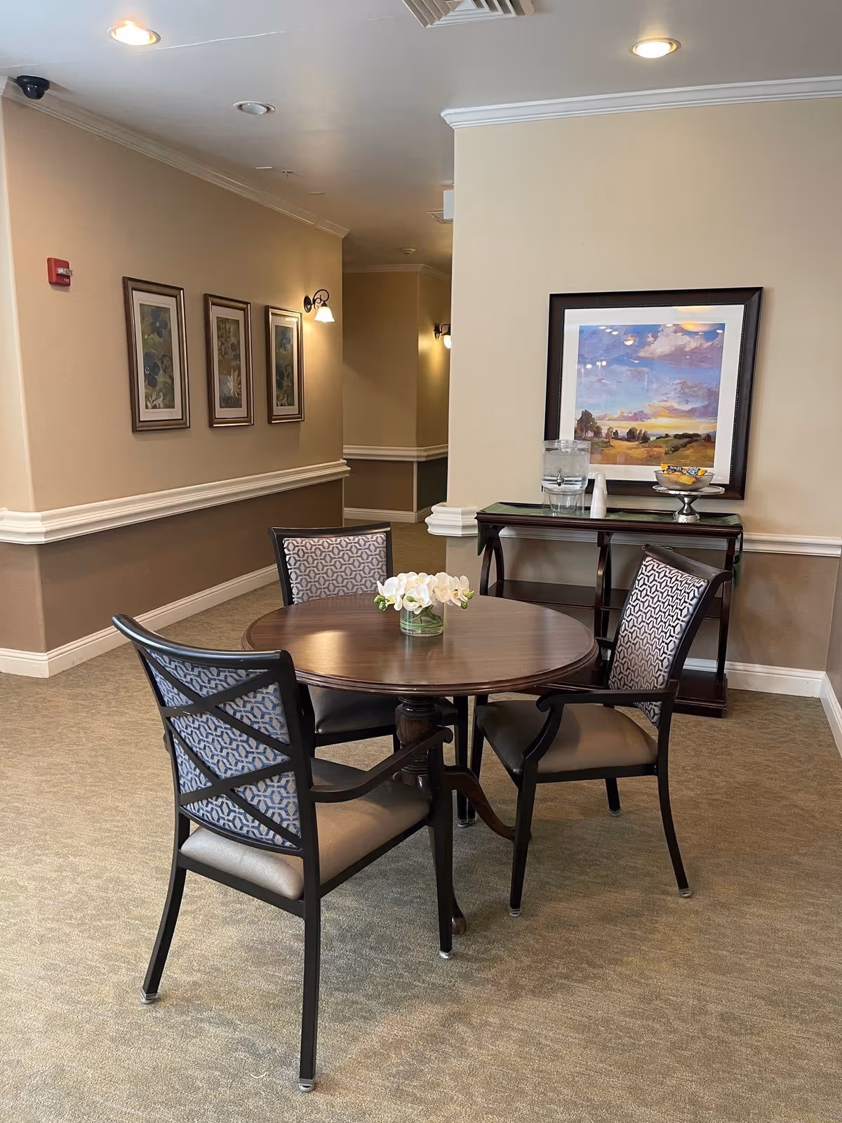 A small round wooden table with a vase of white flowers in the center, surrounded by three cushioned chairs with patterned backs. Behind the table is a wooden console table with a glass water dispenser, a bowl of snacks, and a framed landscape painting on the wall. The room has beige walls with white trim, carpeted floor, and wall-mounted light fixtures.