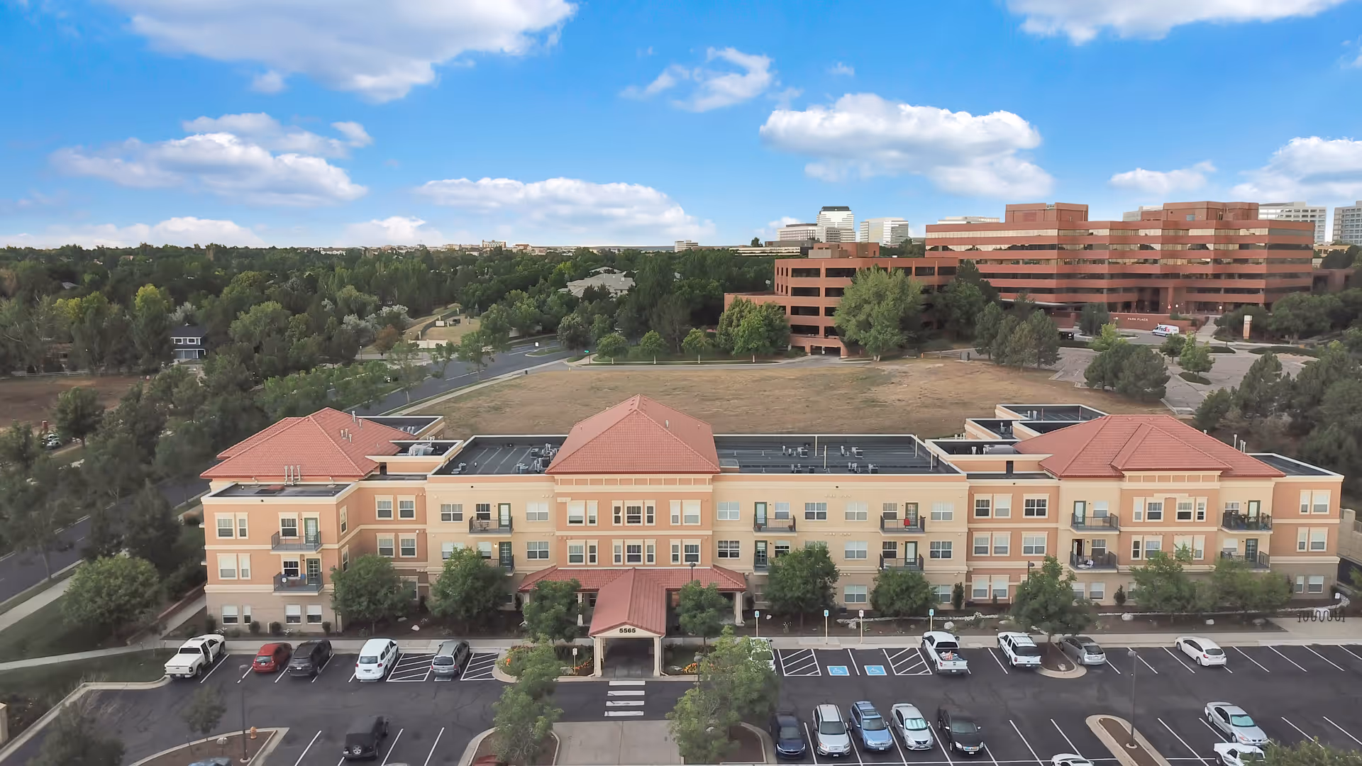 Aerial view of The Inn at Greenwood Village, a three-story residential building with a red-tiled roof and beige exterior. The building is surrounded by trees and a parking lot with several cars parked. In the background, there are office buildings and a partly cloudy blue sky.