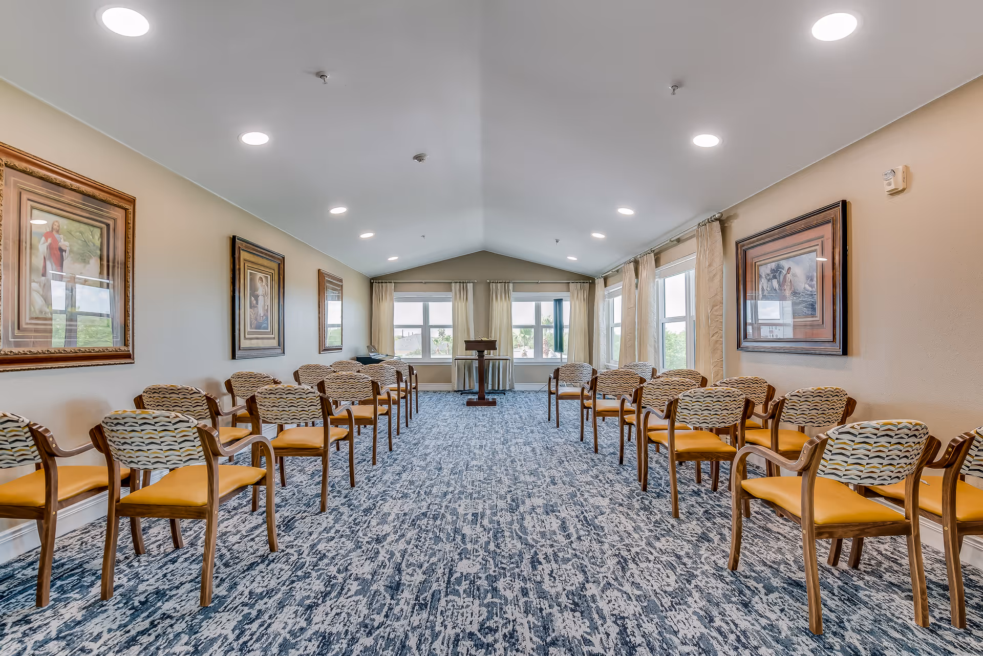 A bright, spacious room with rows of wooden chairs featuring patterned backs and yellow seats arranged facing a wooden podium at the far end. The room has large windows with beige curtains letting in natural light, a blue patterned carpet, and framed artwork on the walls.