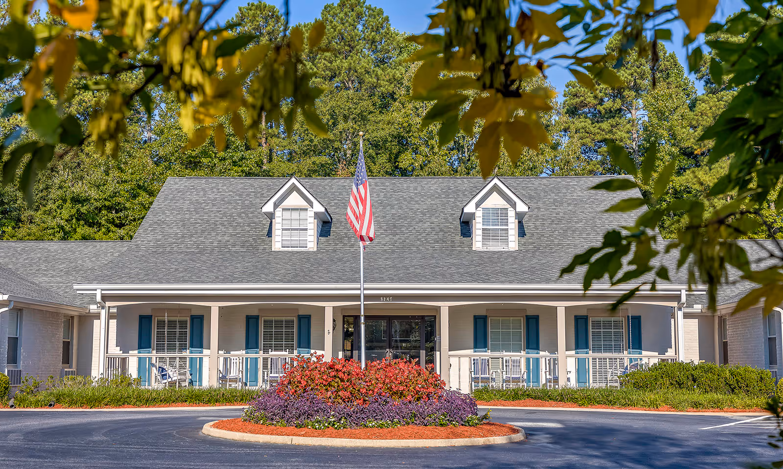 Front exterior view of a single-story senior living facility building with a gray roof, white walls, blue window shutters, and a porch with rocking chairs. An American flag is displayed on a flagpole in front of the building, surrounded by a circular flower bed with red and purple flowers. Trees and greenery are visible in the background.