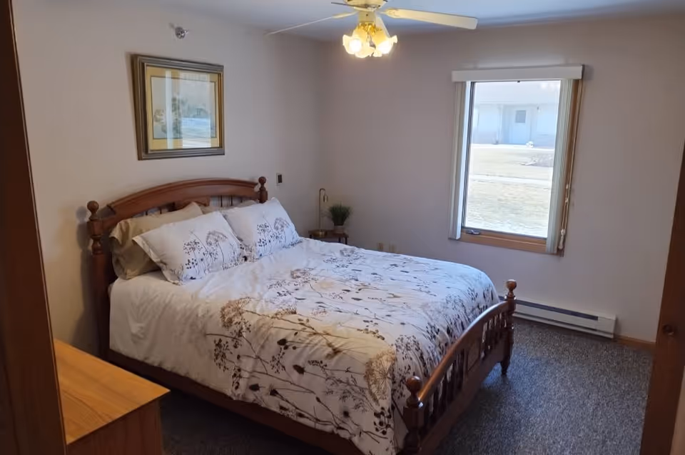 A bedroom with a wooden bed frame and a floral patterned white comforter and pillows. There is a window with vertical blinds letting in natural light, a ceiling fan with lights, a framed picture on the wall above the bed, and a small wooden nightstand with a plant and lamp.