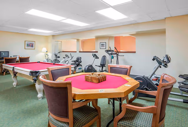 A recreational room with a red felt octagonal game table surrounded by four wooden chairs with cushioned seats. Behind the game table, there is a pool table with red felt. The room also contains several exercise machines including stationary bikes and elliptical trainers. The walls are light-colored, and the floor is carpeted in green. There are framed pictures on the walls and a lamp on a side table near a desk with a computer.