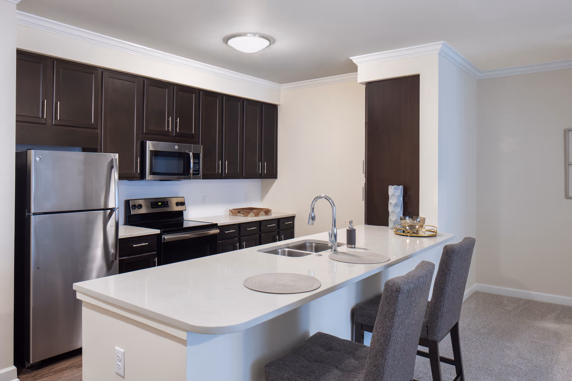 Modern kitchen with dark wood cabinets, stainless steel refrigerator, microwave, and stove. A white countertop island with a built-in sink and two gray upholstered chairs is in the foreground. The kitchen has a clean and minimalist design with neutral wall colors and a ceiling light fixture.