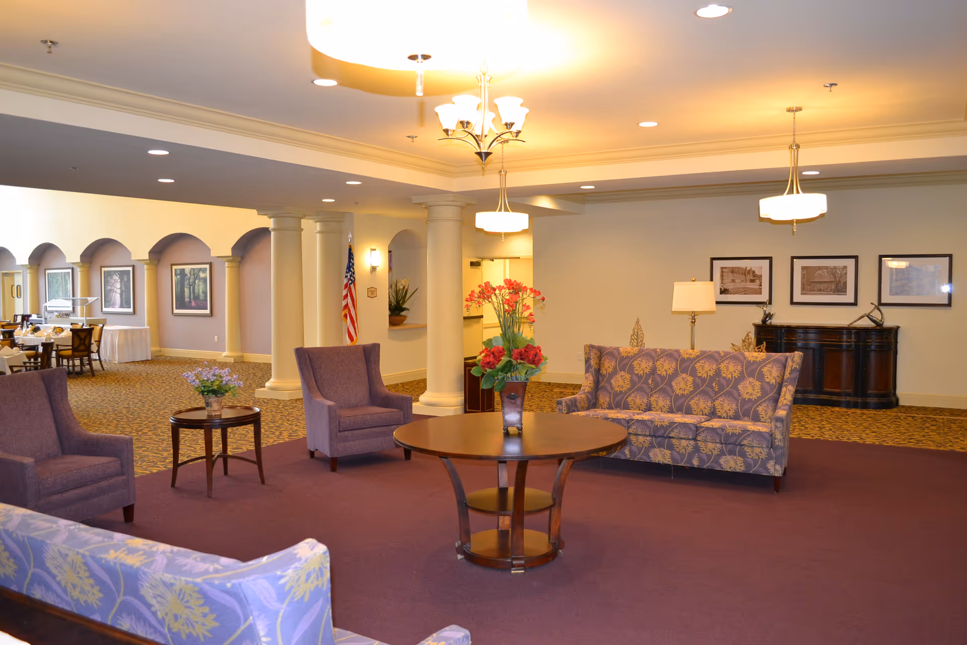A spacious and well-lit common area with purple carpet and floral-patterned sofas and armchairs arranged around a round wooden coffee table with a flower vase. The room features white columns, framed pictures on the walls, and a sideboard with a lamp. In the background, there is a dining area with tables and chairs, and an American flag is visible near the columns.