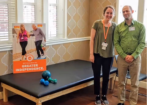 Two staff members standing next to a black padded therapy table in a room with patterned wallpaper and windows with white blinds. On the table are small exercise weights and a poster that reads 'Get on a path to greater independence' with an image of a woman assisting an elderly man with walking.