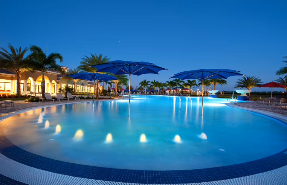 A large, illuminated outdoor swimming pool at dusk with blue umbrellas in the shallow end, surrounded by palm trees and lounge chairs. The building adjacent to the pool is lit warmly, and the sky is clear with a deep blue hue.