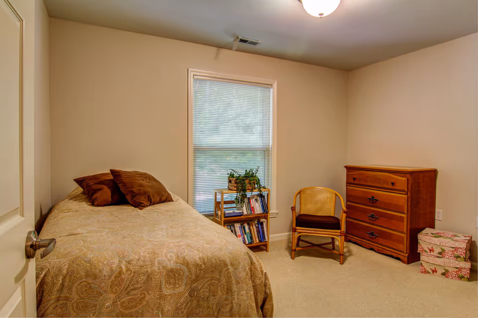 A small bedroom with a single bed covered in a patterned beige bedspread and two brown pillows. Next to the bed is a window with blinds, and below the window is a small wooden bookshelf with books and a potted plant. To the right of the bookshelf is a wooden chair with a woven back and a dark cushion. A wooden dresser with four drawers stands against the right wall, and two floral-patterned storage boxes are on the floor beside it. The walls are painted light beige and the floor is carpeted.
