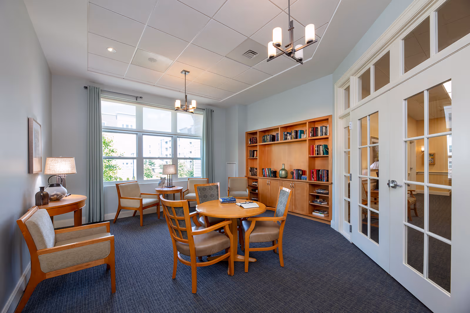 Bright communal sitting room with a round wooden table and chairs, bookshelves, a large window, and glass double doors.