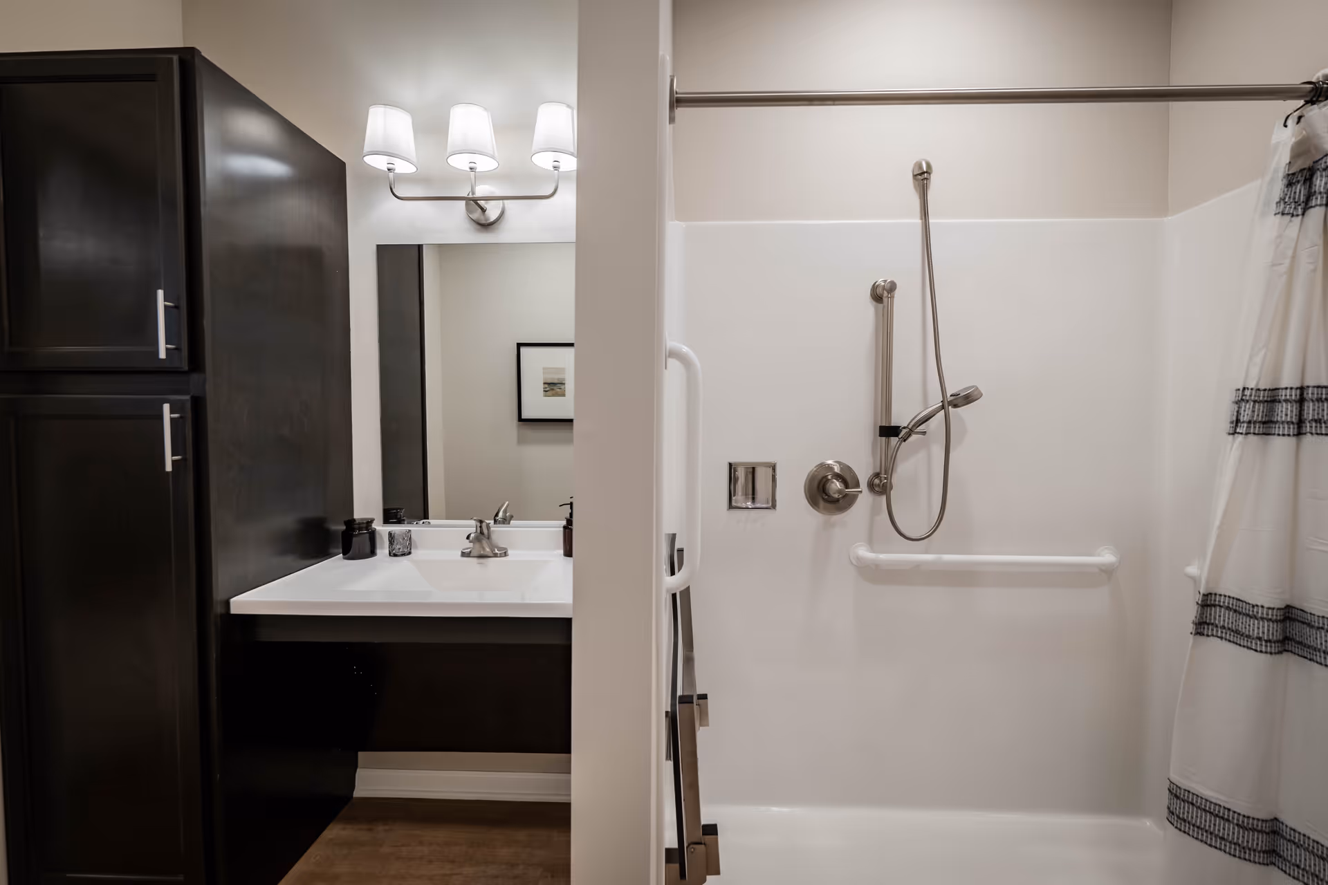 A bathroom with a white sink and dark wood cabinetry on the left side, a large mirror with three light fixtures above it, and a shower area on the right side with a handheld showerhead, grab bars, and a white shower curtain with black stripes.
