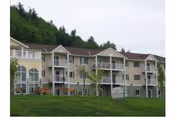 Three-story beige senior living building with multiple balconies facing a grassy lawn and trees in the background.