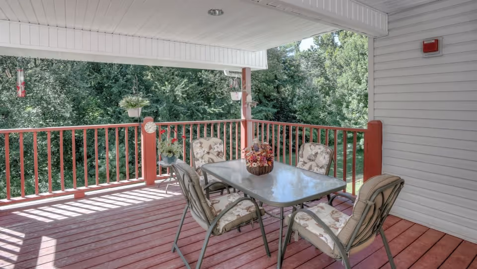 Covered outdoor patio area with a red wooden floor and railing, featuring a glass-top table surrounded by four cushioned chairs with floral patterns. There are hanging plants and bird feeders attached to the railing, and lush green trees are visible in the background.
