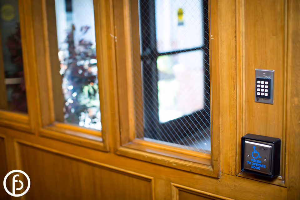Close-up view of a wooden wall panel with a window featuring wire-reinforced glass. Mounted on the wall are an electronic keypad and a blue accessibility button labeled 'Press to operate door' with a wheelchair symbol.