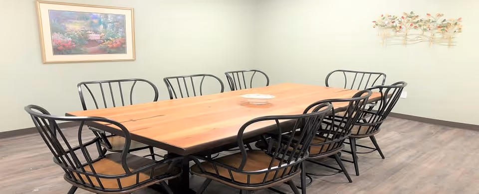 A dining room with a large rectangular wooden table surrounded by eight black metal chairs with wooden seats. The room has light green walls, a framed floral painting on one wall, and a decorative metal wall art piece with flowers on another wall. The floor is a wood-like laminate.