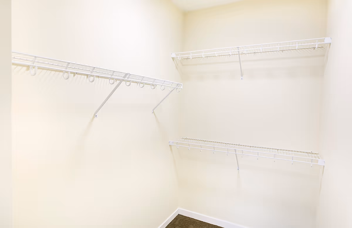 Empty walk-in closet with white wire shelving mounted on cream-colored walls and a small carpeted floor area.