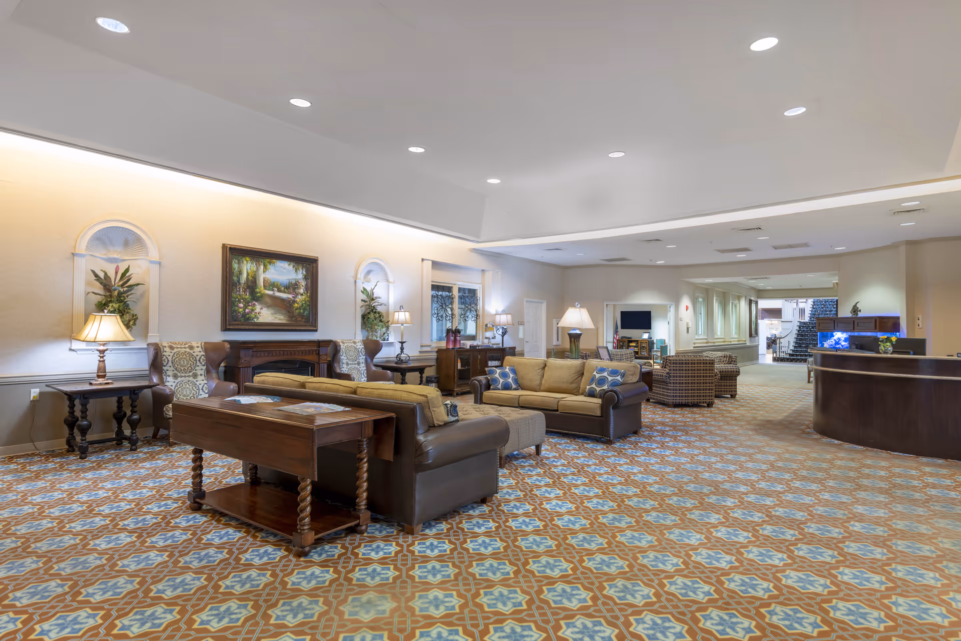 Spacious senior living facility lobby with sofas, armchairs, patterned carpet, lamps, artwork, and a reception desk.