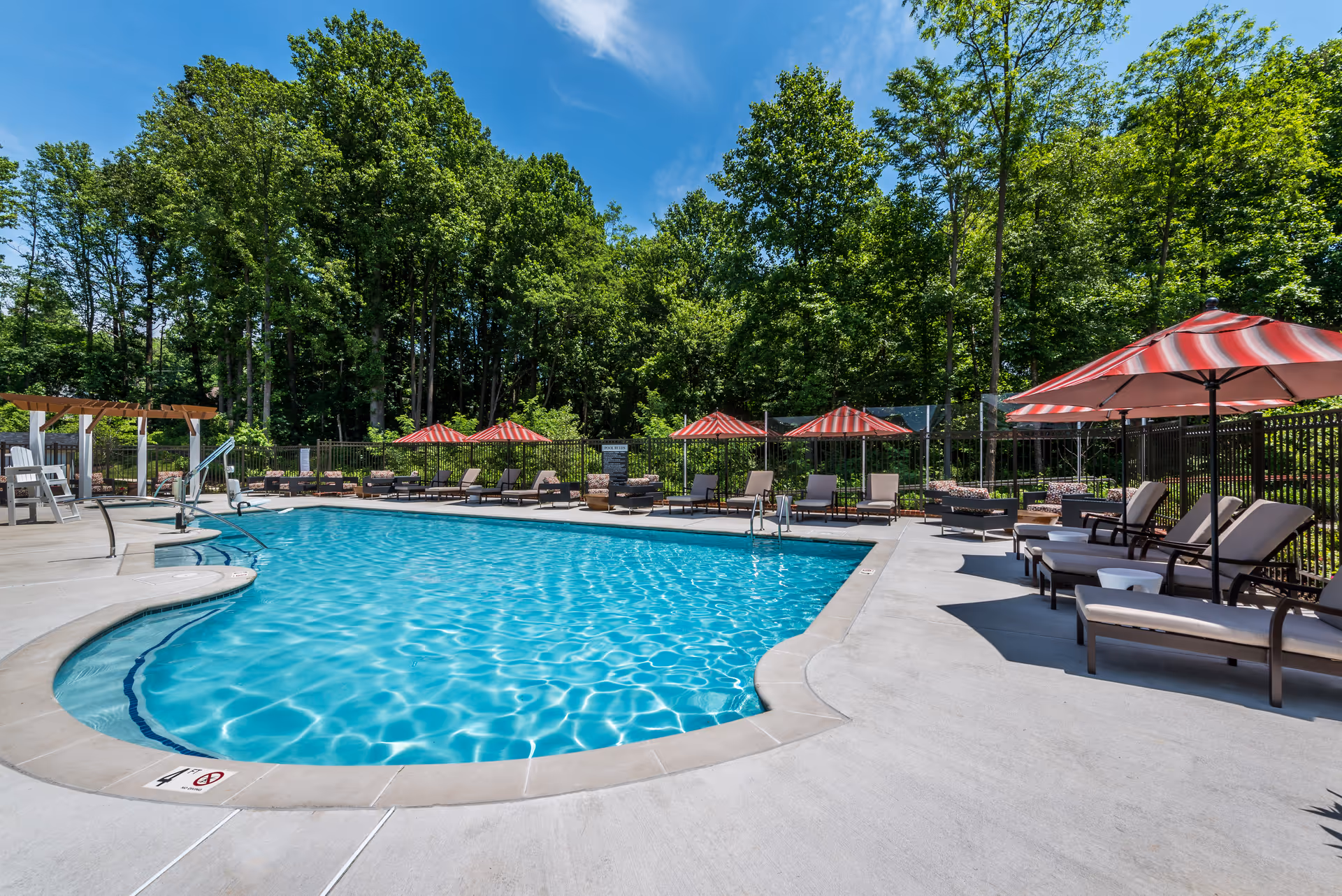 Outdoor swimming pool area with clear blue water, surrounded by lounge chairs and red-and-white striped umbrellas. The pool area is bordered by a fence and lush green trees under a bright blue sky with some clouds.