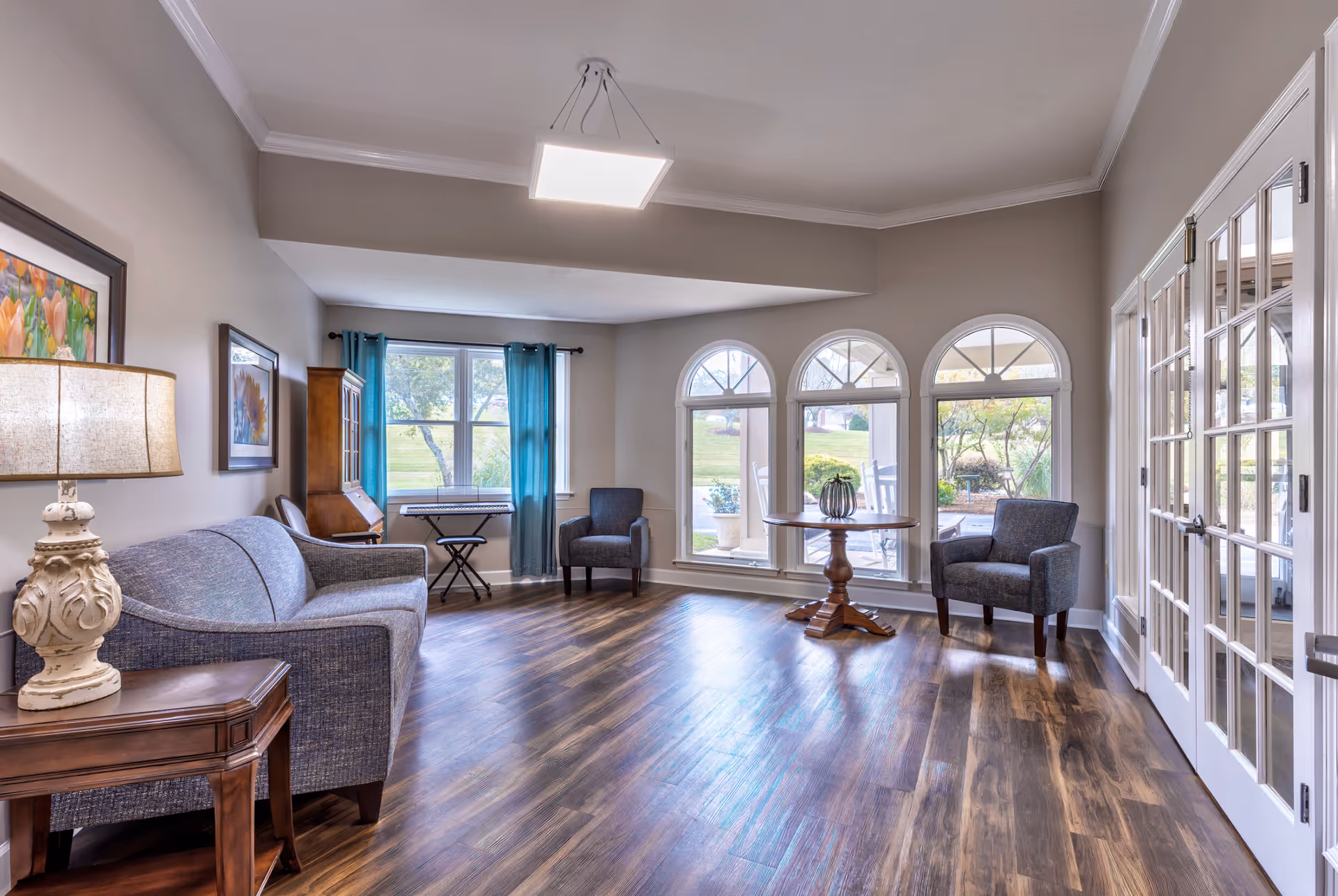 A bright and spacious living room area with wooden flooring, a blue upholstered sofa, two gray armchairs, a round wooden table with a decorative item, and large arched windows letting in natural light. There is a lamp on a wooden side table, framed floral artwork on the walls, and a keyboard near the window with blue curtains. French doors are visible on the right side leading to another room or outdoor area.