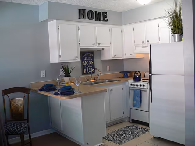 Compact white kitchen with cabinets, refrigerator, stove, sink, and a breakfast bar decorated with place settings and 'HOME' letters above the cabinets.