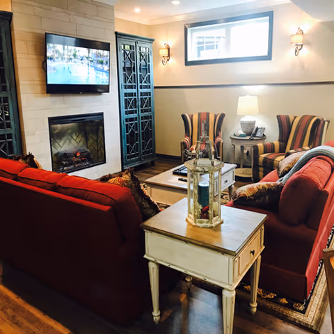 Cozy living room with red sofas, striped armchairs, a fireplace under a wall-mounted TV, and a small side table with a decorative lantern.