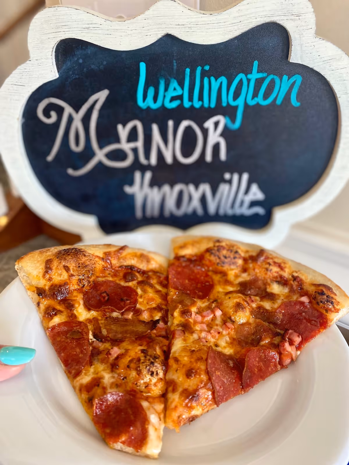 Two slices of pepperoni pizza on a white plate with a small decorative chalkboard sign behind them that reads 'Wellington Manor Knoxville'.