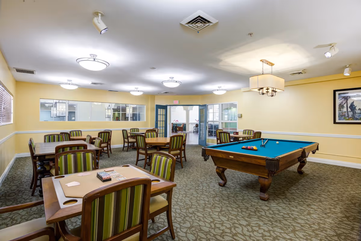 A spacious recreational room with several wooden tables and chairs featuring green striped upholstery. One table has playing cards and poker chips set up. There is a pool table with a blue felt surface and billiard balls arranged for a game. The room has yellow walls, carpeted flooring, multiple ceiling lights, and a framed picture on the wall. Double glass doors lead to another area.