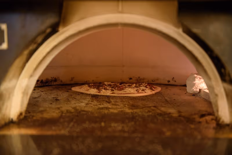 A pizza baking inside a traditional stone or brick pizza oven with a visible flame on the right side.