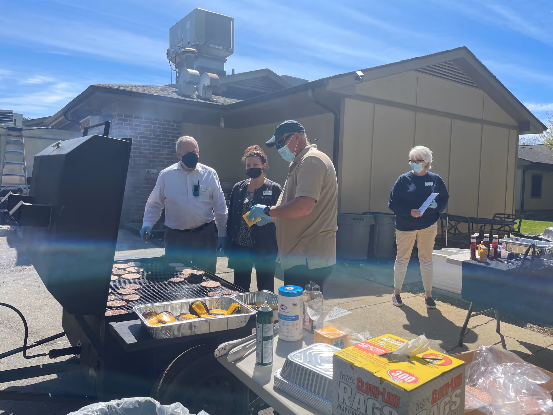 Several masked people stand outdoors around a large barbecue grill cooking burgers with condiments and supplies on nearby tables in front of a facility building.