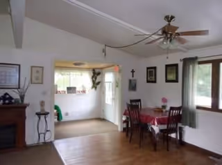 Interior view of a dining area with a wooden table covered with a red and white tablecloth and four chairs. There is a ceiling fan with lights above the table. The room has white walls with framed pictures and a cross hanging. A window with green curtains lets in natural light. Adjacent to the dining area is a small entryway with a door and a window, decorated with a wall-mounted sculpture and a sign that reads 'ENTER HERE'.