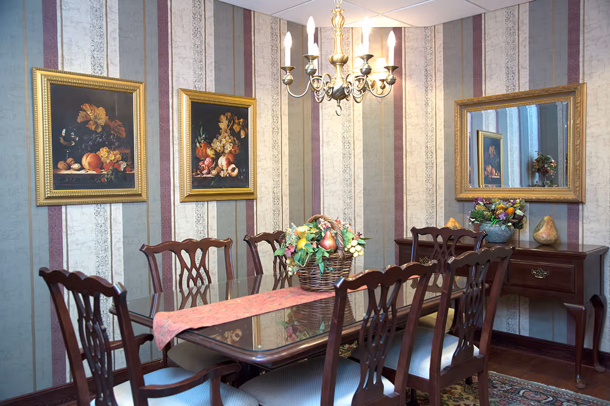A traditional dining room with a polished wooden table and six matching chairs. The table has a pink runner and a centerpiece basket filled with artificial fruit and flowers. The walls are decorated with vertical striped wallpaper in shades of gray, white, and maroon, and adorned with two framed still life paintings of fruit and a large gold-framed mirror. A chandelier with multiple candle-style lights hangs above the table. A wooden side table with a floral arrangement and decorative gourds is positioned against the wall beneath the mirror.