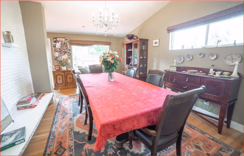 A dining room with a long table covered by a red tablecloth and surrounded by six dark wooden chairs with leather seats. A vase with flowers is placed in the center of the table. The room has a wooden floor with a patterned rug underneath the table. There is a wooden cabinet with glass doors and a sideboard displaying decorative plates and cups. A window with a valance lets in natural light, and a chandelier hangs from the ceiling. The walls are painted beige, and there is a small seating area with a couch near the window.
