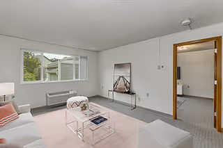 A bright living room with a large window showing greenery outside. The room features a white sofa with pink and white pillows, a pink rug, a glass coffee table with magazines, a small ottoman, and a framed picture of a pier on the wall. There is an open doorway leading to another room with a TV visible.